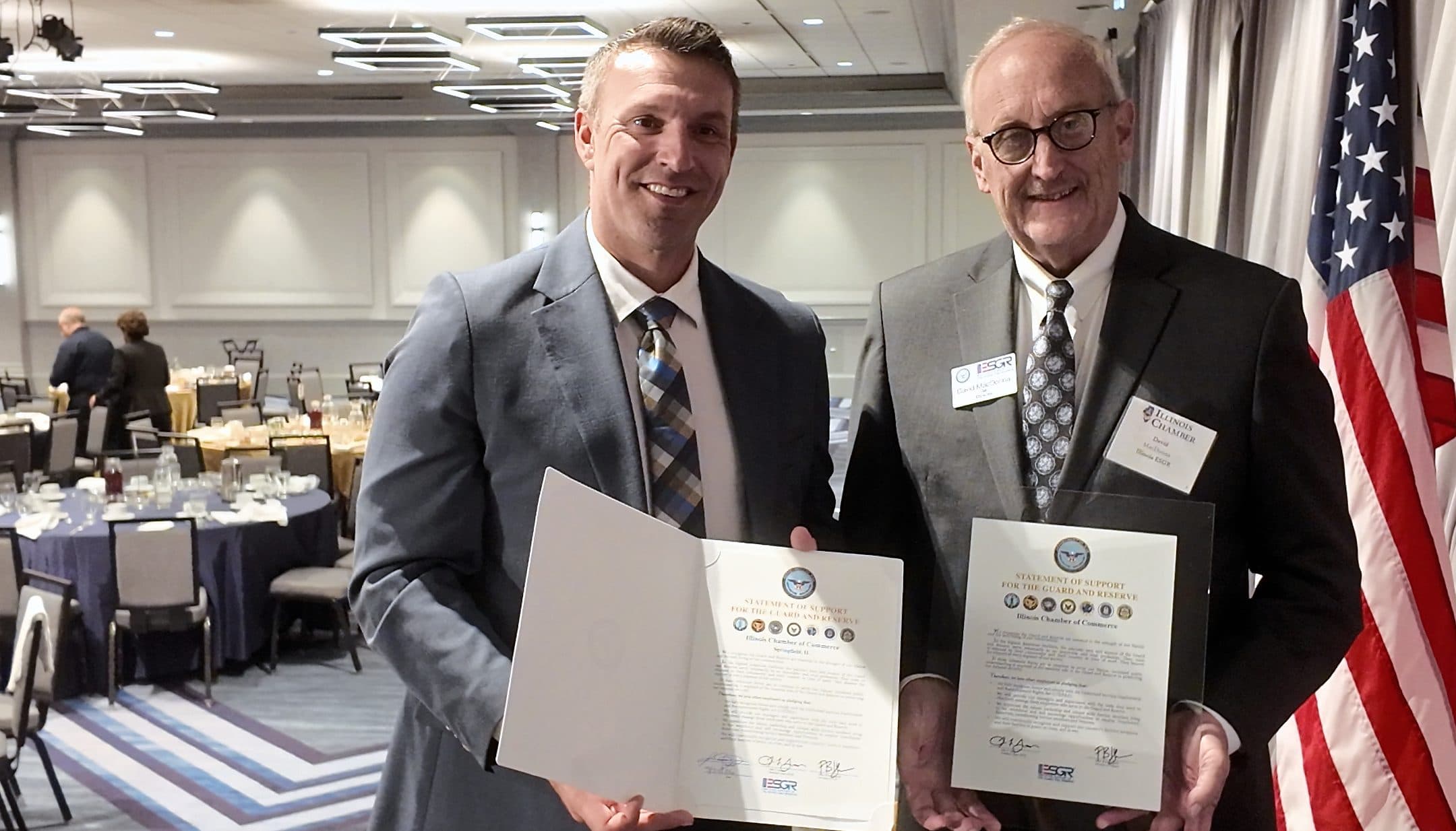 Two men hold signed certificates beside an American flag.
