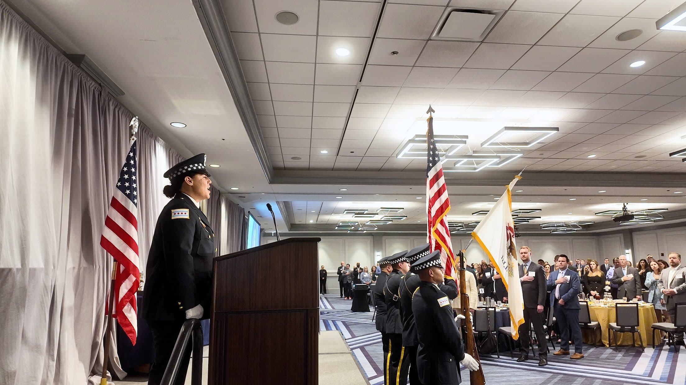 A uniformed officer stands at the podium as the honor guard holds the flags.
