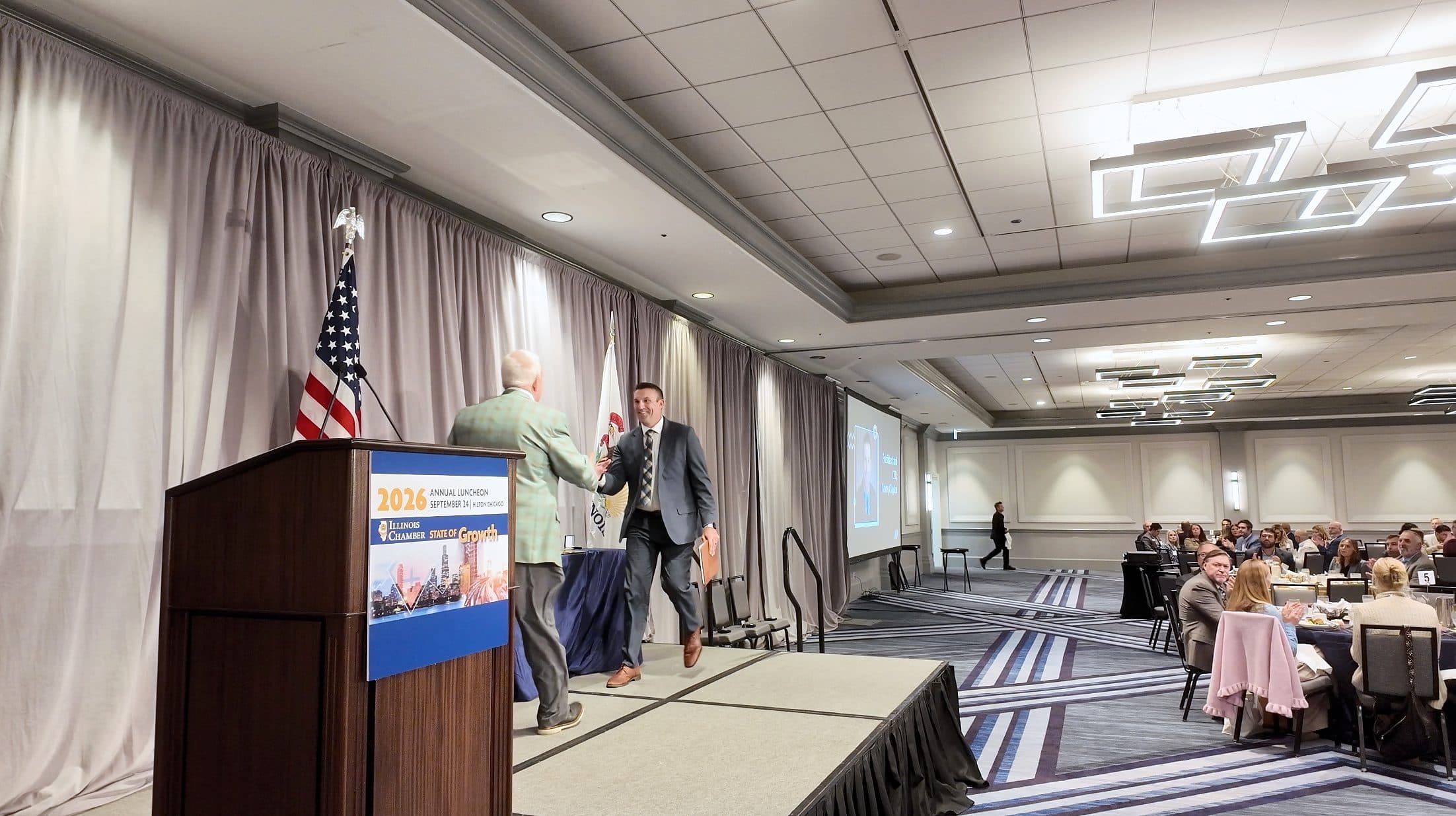 Two men shake hands on stage beside the podium.